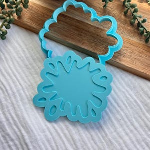 May include: A bright cyan plastic cookie cutter featuring a two-part design. The top section is a scalloped cloud outline, while the bottom section is a solid, irregular starburst or splash shape with several internal cutouts for detailed impressions.