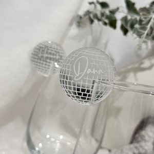 May include: Two clear cocktail glasses with a silver disco ball-shaped drink stirrer. The word "Dana" is etched on the disco ball. The background is a soft, blurred white.