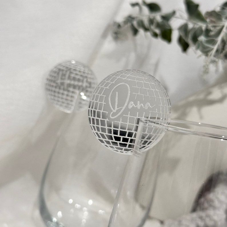 May include: Two clear cocktail glasses with a silver disco ball-shaped drink stirrer. The word "Dana" is etched on the disco ball. The background is a soft, blurred white.