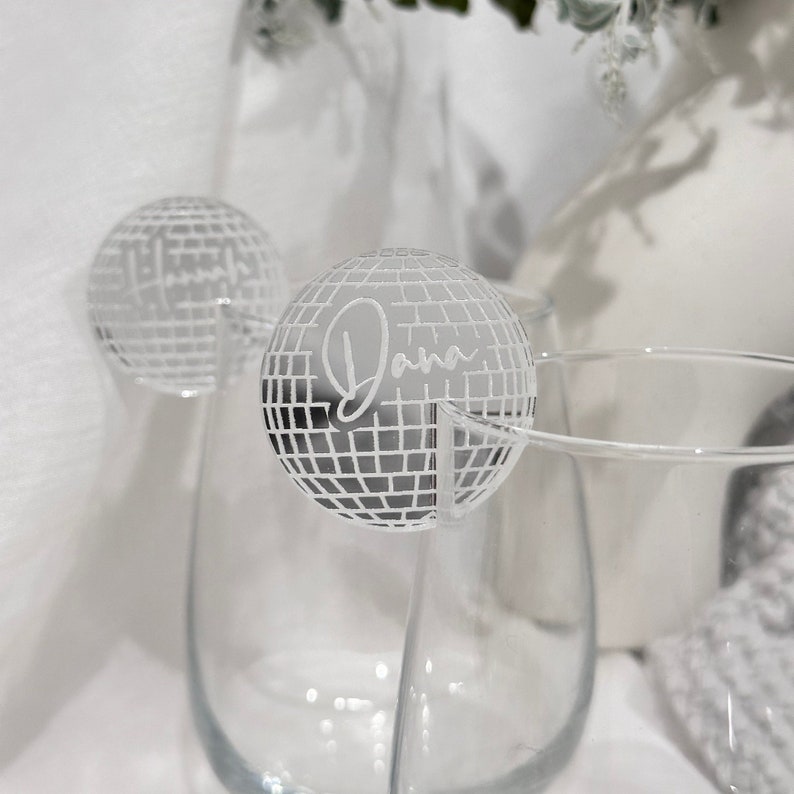 May include: Clear glass wine glasses with a decorative disco ball-shaped name tag that reads "Dana" in a cursive font. The disco ball is silver and has a grid pattern. The background is white.
