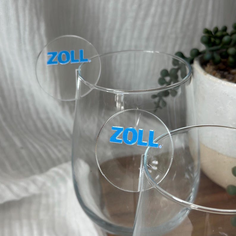May include: Clear wine glasses with round, transparent drink charms. Each charm has the word "ZOLL" printed in blue. The glasses are partially filled, and the background includes a white cloth and a potted plant.