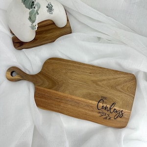 May include: A rectangular wooden cutting board with a handle and a hole for hanging. The board has a natural wood grain and the text "The Conleys" is engraved on the lower right corner with a leaf design.