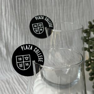 May include: Two clear wine glasses with black circular Plaza College logo coasters. The coasters have the words "Plaza College" in a circular pattern, with the numbers "19" and "16" and a crown symbol. The background is a soft, textured white.