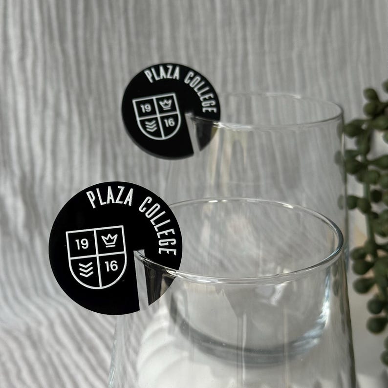 May include: Two clear wine glasses with black circular Plaza College logo coasters. The coasters have the words "Plaza College" in a circular pattern, with the numbers "19" and "16" and a crown symbol. The background is a soft, textured white.