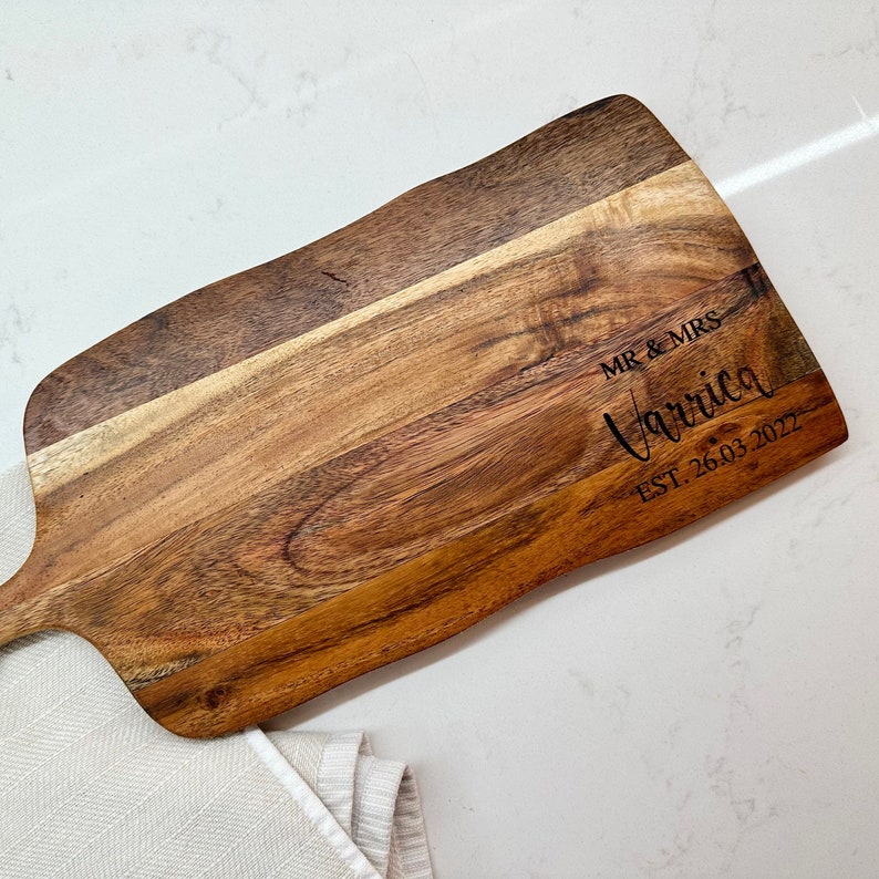 May include: A wooden serving board with a handle, featuring a natural wood grain pattern. The board is engraved with "MR & MRS Varriga EST. 26.03.2022" in a stylish font. The board rests on a white surface with a folded white cloth.