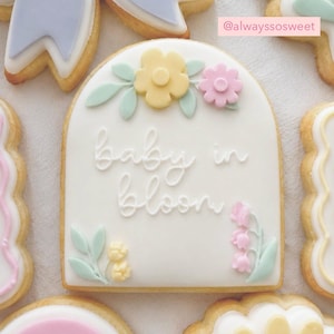 Może przedstawiać: Ozdobione ciastko maślane z białym lukrem i napisem "baby in bloom" kursywą. Ciastko jest ozdobione żółtymi i różowymi kwiatami oraz zielonymi liśćmi. Inne zdobione ciastka otaczają główne.