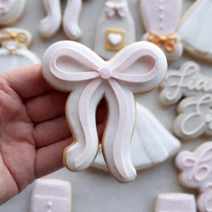 Op de afbeelding: Een witte koek in de vorm van een strik met roze lintdetails. De koek heeft een lichtbruine rand en wordt in een hand gehouden. Andere koekjes op de achtergrond zijn een trouwjurk, een pak en een champagnefles.