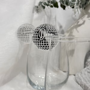 May include: Clear stemless wine glasses with a disco ball-shaped drink charm. The charm is black and white with the name "Dana" in a cursive font. The background is white with a hint of greenery.