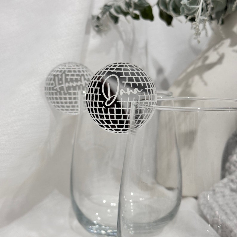 May include: Clear stemless wine glasses with a disco ball-shaped drink charm. The charm is black and white with the name "Dana" in a cursive font. The background is white with a hint of greenery.