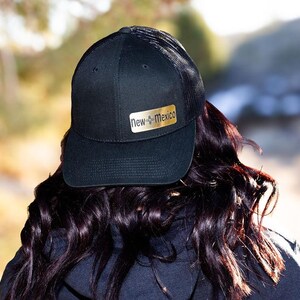 May include: Black baseball cap with a mesh back and a gold rectangular patch that reads "New Mexico". The cap is worn by a person with dark hair. The background is blurred, with hints of green and yellow.