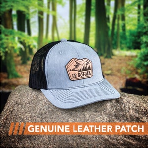 Puede incluir: Una gorra de camionero gris y negra con un parche de cuero marr&oacute;n que dice "I'd Rather Be Outside". El parche presenta una cordillera y &aacute;rboles.