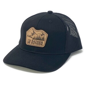 Puede incluir: Gorra de camionero negra con un parche de cuero marr&oacute;n que dice "I'd Rather Be Outside" y presenta una silueta de monta&ntilde;a.