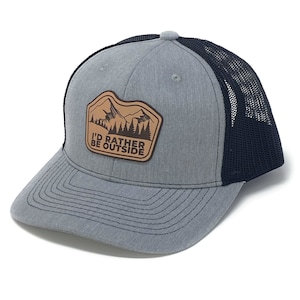 Puede incluir: Una gorra de camionero gris y negra con un parche de cuero marr&oacute;n que dice "I'd Rather Be Outside". El parche presenta una silueta de monta&ntilde;as y &aacute;rboles.