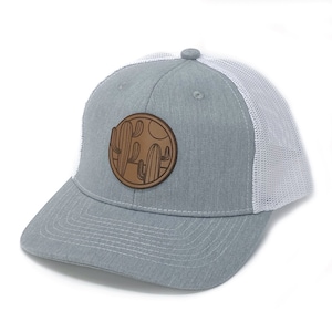 May include: A gray and white trucker hat with a brown leather patch featuring a cactus design. The patch is circular and has a white background with brown outlines of cacti and a sun.