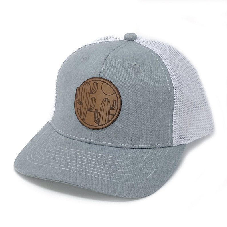 Puede incluir: Una gorra de camionero gris y blanca con un parche de cuero marr&oacute;n con un dise&ntilde;o de cactus. El parche es circular y tiene un fondo blanco con contornos marrones de cactus y un sol.