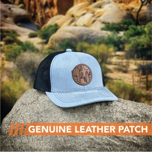 Puede incluir: Una gorra de camionero azul claro y negra con un parche de cuero marr&oacute;n que presenta un dise&ntilde;o de cactus. El parche tiene el texto "Yucca" en un c&iacute;rculo alrededor del cactus.