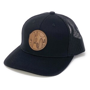 Puede incluir: Una gorra de camionero negra con un parche de cuero marr&oacute;n que presenta una silueta de cactus y una luna. La gorra tiene una parte trasera de malla.