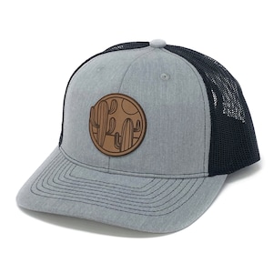 Puede incluir: Una gorra de camionero gris y negra con un parche de cuero marr&oacute;n con un dise&ntilde;o de cactus.