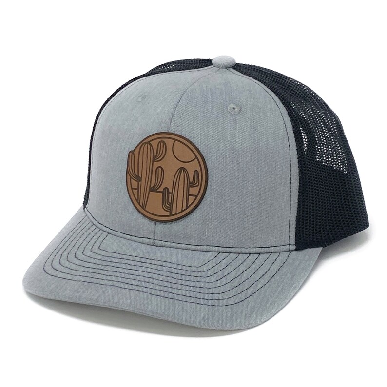 Puede incluir: Una gorra de camionero gris y negra con un parche de cuero marr&oacute;n con un dise&ntilde;o de cactus.