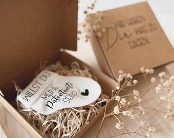 Könnte beinhalten: Eine kleine weiße Socke mit dem Text "Willst du meine Patenante sein?" und einem schwarzen Herzen. Die Socke befindet sich in einer braunen Box mit zerkleinertem Papier.