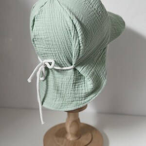 May include: A light green cotton sun hat with a wide brim and a white drawstring tie. The hat is displayed on a wooden stand.