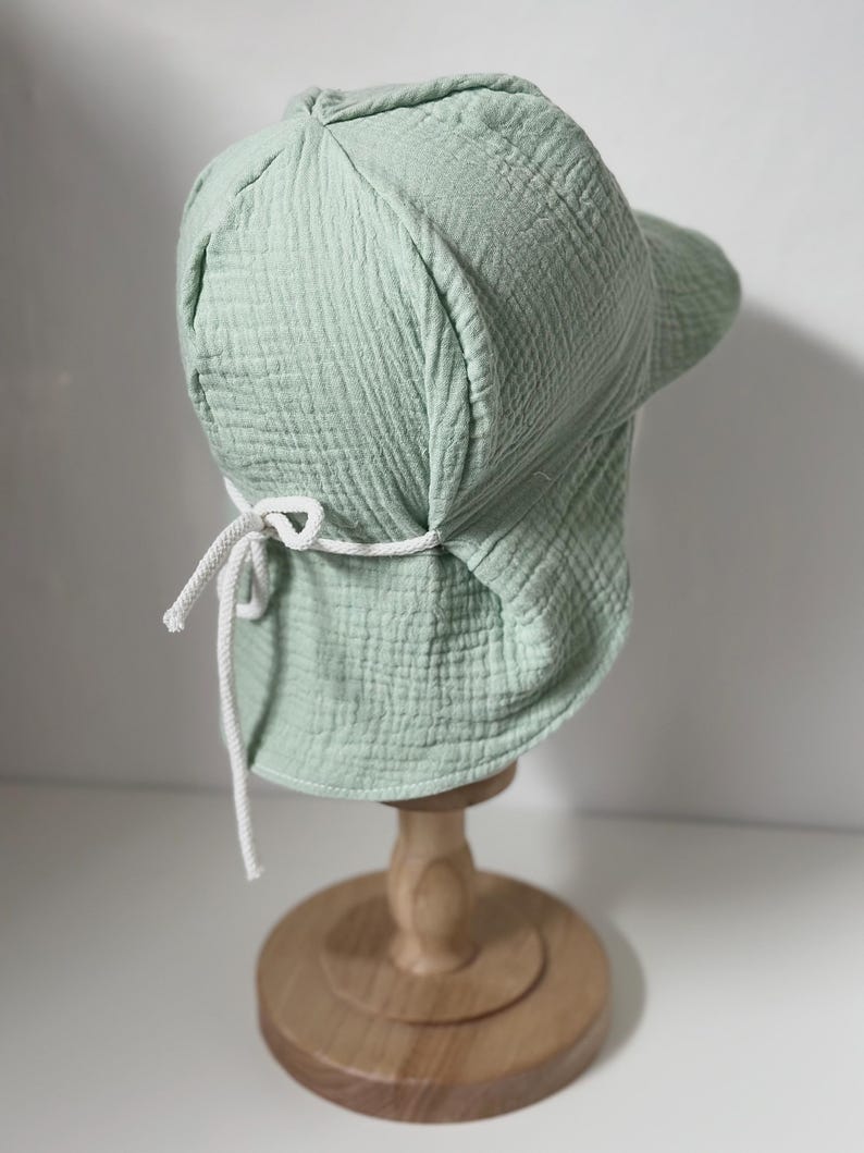 May include: A light green cotton sun hat with a wide brim and a white drawstring tie. The hat is displayed on a wooden stand.