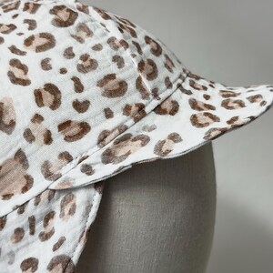 May include: A white sun hat with a brown and white leopard print pattern. The hat has a wide brim and a flap that can be pulled down to protect the neck.