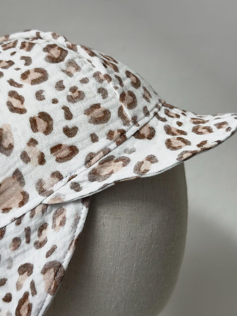 May include: A white sun hat with a brown and white leopard print pattern. The hat has a wide brim and a flap that can be pulled down to protect the neck.