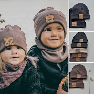 Könnte beinhalten: Zwei Kinder tragen braune Strickmützen mit den Worten "little sis" und "big bro" darauf. Die Kinder tragen passende braune Strickschals. Das Bild zeigt auch eine Auswahl an Mützen in verschiedenen Farbkombinationen mit dem gleichen Text.