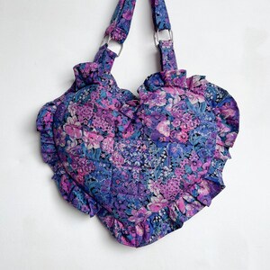 May include: A heart-shaped handbag with a floral print in shades of purple, pink, and blue. The bag has a ruffled edge and two fabric straps.