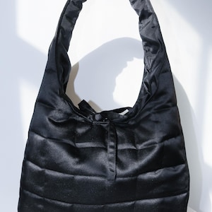 May include: Black quilted satin tote bag with a rounded handle. The bag features a rectangular shape with horizontal stitching and a tie closure. The bag is made of a shiny, smooth material.