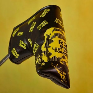 Puede incluir: Cubierta negra para la cabeza del palo de golf con un diseño pixelado amarillo de un personaje de videojuego. La cubierta tiene el texto "RIP" y "GAME OVER" repetido.