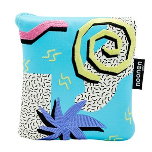 May include: A blue pouch with a 90s-inspired geometric pattern, featuring a yellow spiral, pink and black lines, and a purple palm tree. The pouch has a black tag with the text "noonan golf" on it.