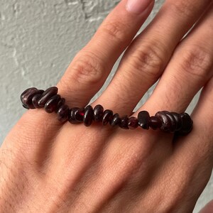 May include: A dark red beaded bracelet. The bracelet is made of small, irregularly shaped beads. The beads are strung together on a flexible cord. The bracelet is shown on a person's hand.