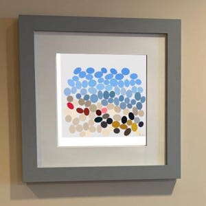 May include: A square framed wall art piece featuring a grey frame and a white mat. The artwork displays a pattern of numerous small, rounded oval shapes in various colours including light blue, beige, red, dark brown, black, and mustard yellow.
