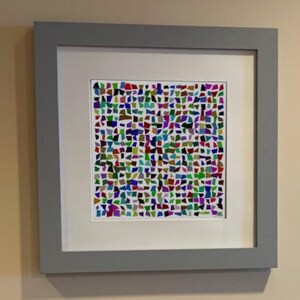 Può includere: Un'opera d'arte quadrata incorniciata con un passe-partout bianco e una cornice grigia. L'immagine centrale consiste in una disposizione a griglia di numerose piccole forme di carta irregolari e multicolori nei toni del rosso, blu, verde, viola e marrone.