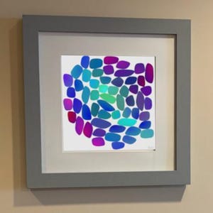 May include: A square framed wall art piece featuring a white background with a central cluster of rounded, pebble-like shapes in gradient shades of blue, teal, purple, and magenta. The artwork is enclosed in a thick, matte grey square frame.