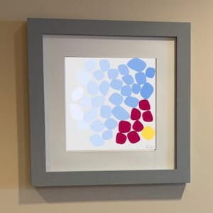 Può includere: Una stampa quadrata incorniciata che presenta una composizione astratta di forme arrotondate in tonalità azzurro, bianco e rosa scuro, con un singolo cerchio giallo posizionato nell'angolo in basso a destra, su uno sfondo grigio chiaro.