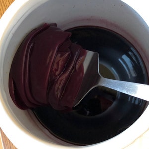 May include: A white container with a dark purple substance inside. A silver spoon is partially submerged in the substance.