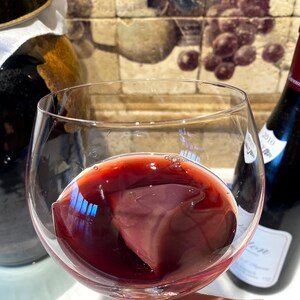 May include: A glass of red wine with a red wine ice cube in it. The wine is in a clear glass with a stem. The glass is being held by a hand.