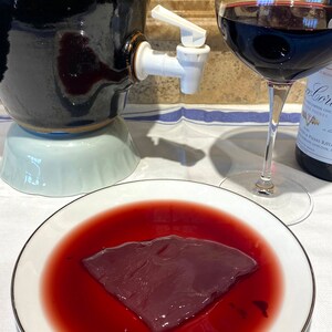 May include: A white ceramic plate with a dark red liquid and a piece of raw meat in the center. The meat is a dark red color and has a slightly textured surface. The liquid is a deep red color and appears to be wine.