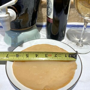 Op de afbeelding: Een ronde, lichtbruine kombucha SCOBY op een wit bord met een zwart en geel meetlint dat 17,8 cm breed is.