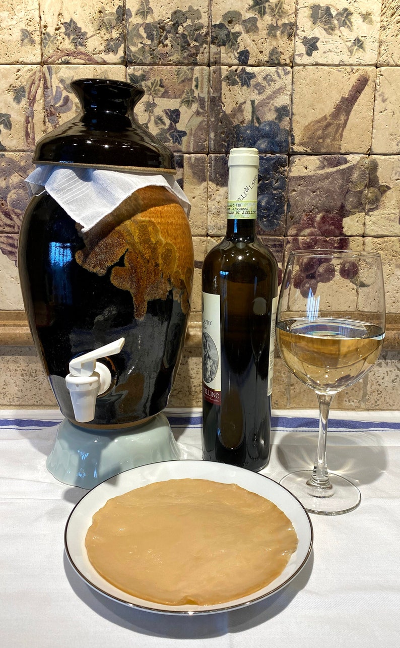 Op de afbeelding: Een keramische pot met een kraan, een fles witte wijn en een glas witte wijn op een wit tafelkleed. De pot is bedekt met een witte doek. De fles heeft een etiket met de tekst "Villoresi" en "Vino Bianco di Antica Tradizione".
