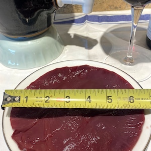 May include: A close-up of a raw, red, round piece of meat on a white plate with a gold trim. A yellow measuring tape is placed on top of the meat, showing a measurement of 6 inches.