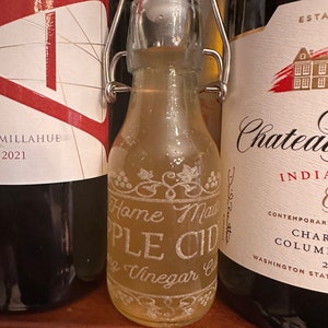 May include: A clear glass bottle with a metal swing top lid filled with a light yellow liquid. The bottle has a white label with the text "Home Made Apple Cider Vinegar" etched into the glass.