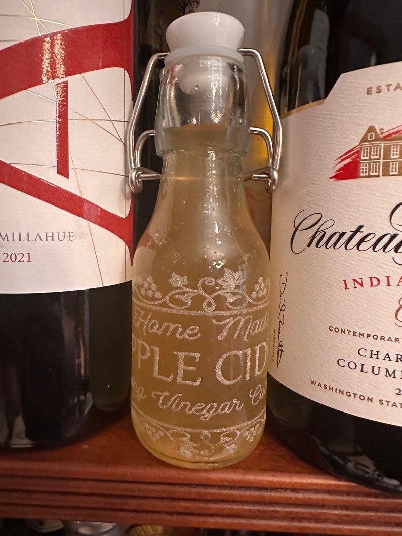 May include: A clear glass bottle with a metal swing top lid filled with a light yellow liquid. The bottle has a white label with the text "Home Made Apple Cider Vinegar" etched into the glass.