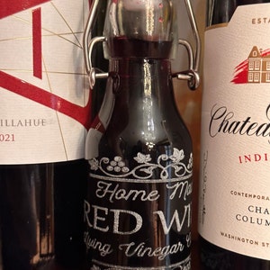 May include: A small glass bottle with a silver metal clasp and a black label that reads "Home Made Red Wine Vinegar". The bottle is filled with a dark red liquid.