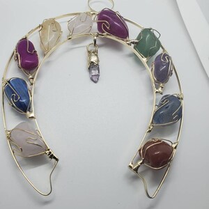 May include: A gold-colored metal horseshoe-shaped pendant with various colorful gemstones. The gemstones include pink, purple, blue, green, and brown stones. A small crystal pendant hangs from the center.