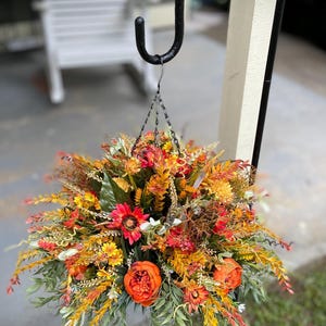 K&ouml;nnte beinhalten: Ein h&auml;ngendes Blumenarrangement mit k&uuml;nstlichen Herbstblumen und Blattwerk in Orange-, Rot-, Gelb- und Gr&uuml;nt&ouml;nen. Das Bouquet enth&auml;lt k&uuml;nstliche Pfingstrosen, G&auml;nsebl&uuml;mchen und verschiedene Gr&auml;ser und h&auml;ngt an einem schwarzen Metallhaken.