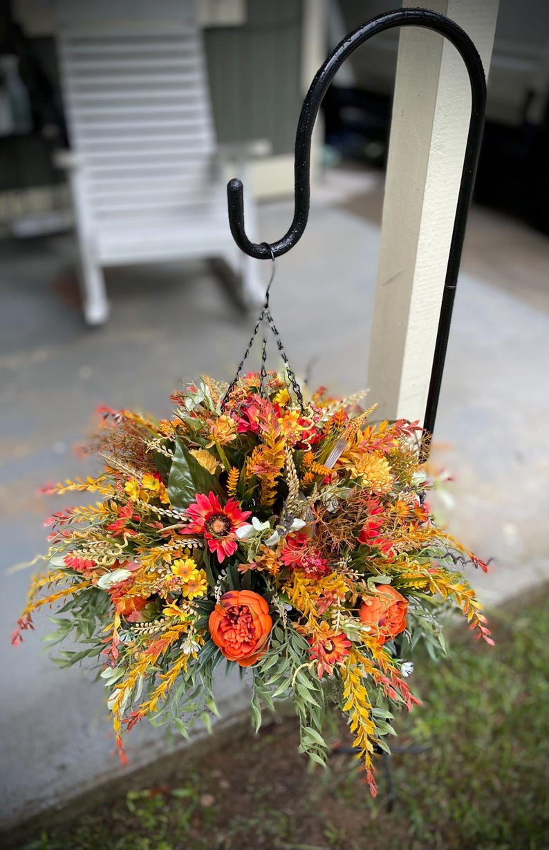 K&ouml;nnte beinhalten: Ein h&auml;ngendes Blumenarrangement mit k&uuml;nstlichen Herbstblumen und Blattwerk in Orange-, Rot-, Gelb- und Gr&uuml;nt&ouml;nen. Das Bouquet enth&auml;lt k&uuml;nstliche Pfingstrosen, G&auml;nsebl&uuml;mchen und verschiedene Gr&auml;ser und h&auml;ngt an einem schwarzen Metallhaken.